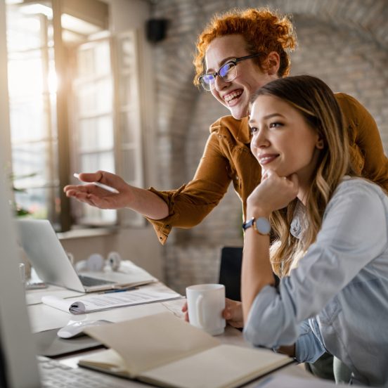 Happy female entrepreneurs reading an e-mail on computer while working together in the office. Focus is on redhead woman.